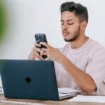 Thoughtful ethnic man browsing smartphone and working on laptop